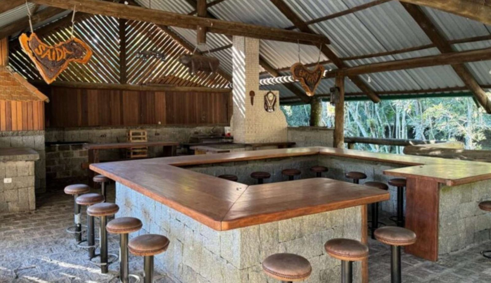 Bar rústico com balcão em pedra e madeira e banquetas, aberto para a mata